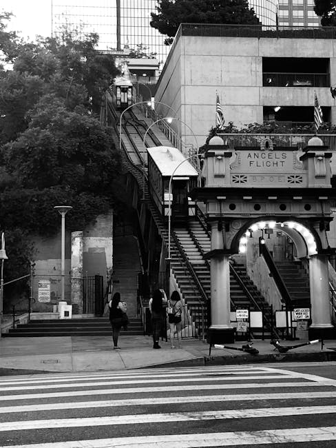 Black and white photograph of an urban street scene showing a group of three people standing on the pavement near a staircase that leads up to an elevated walkway or building entrance. The staircase h