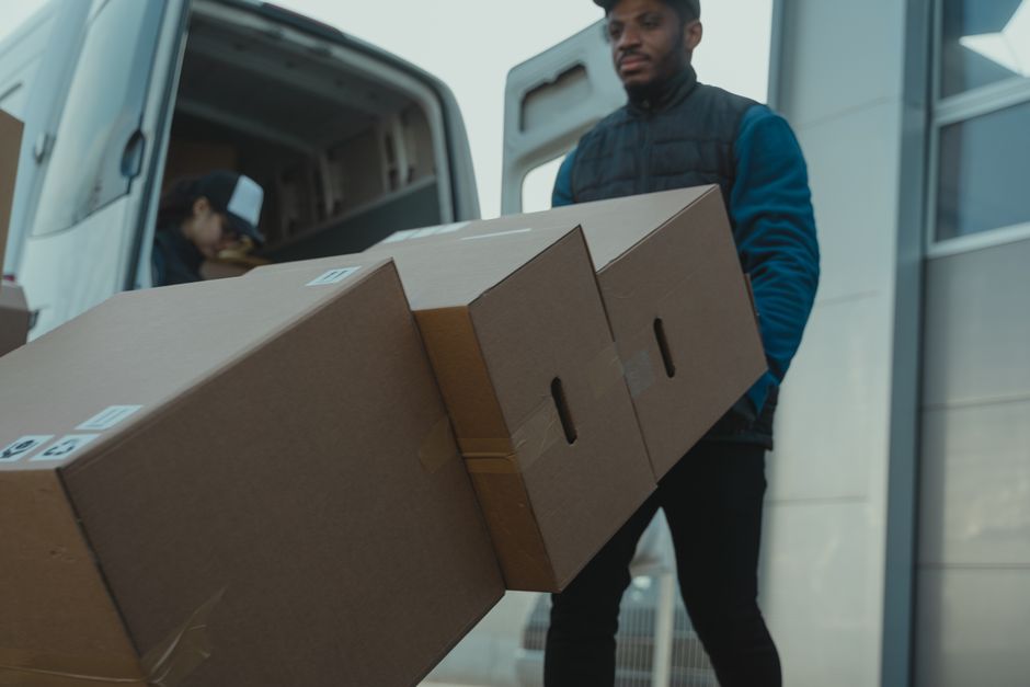 A man dressed in a blue jacket and dark pants is loading multiple medium-sized cardboard boxes, sealed with packing tape and labeled, onto a flat surface outside a white van parked near a modern build