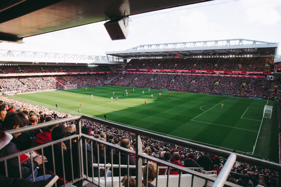 Inside a large football stadium with a well-maintained green pitch and clearly marked white lines, surrounded by multiple tiers of red and white seating sections with the Emirates logo visible on the 