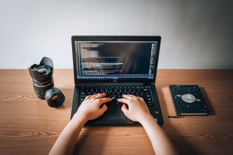 A close-up image of a person's hand using a keyboard on a silver laptop placed on a wooden surface. The laptop screen displays lines of code with syntax highlighting, indicating programming activity.